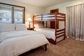 A secondary bedroom at the Ranch at Steamboat RA219 features a queen-sized bed, a wooden bunk bed, a nightstand with a lamp, a window with blinds, and striped curtains against a light-colored wall.