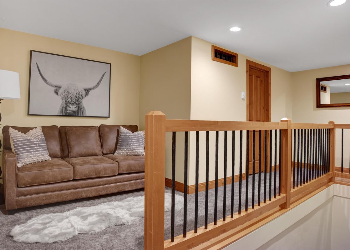 A cozy living area features a brown leather sofa, a floor lamp, and a large black and white art print of a highland cow, with a wooden and metal railing in the foreground.