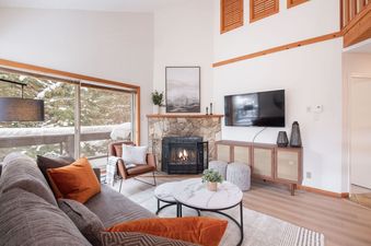 Cozy living room in Foxglove 1 featuring a stone fireplace, flat-screen TV, and large windows looking out onto a snowy pine landscape.