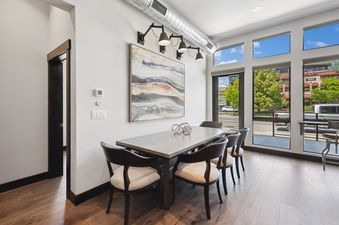 A modern dining area in Downtown Gem features a rectangular table with six chairs, an abstract painting, and large windows with an urban view.