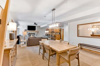An inviting open-concept living and dining area at Christie Club 306 features a stone fireplace, a large TV, a brown sofa, a dining table with six chairs, and a well-appointed kitchen with a breakfast bar.