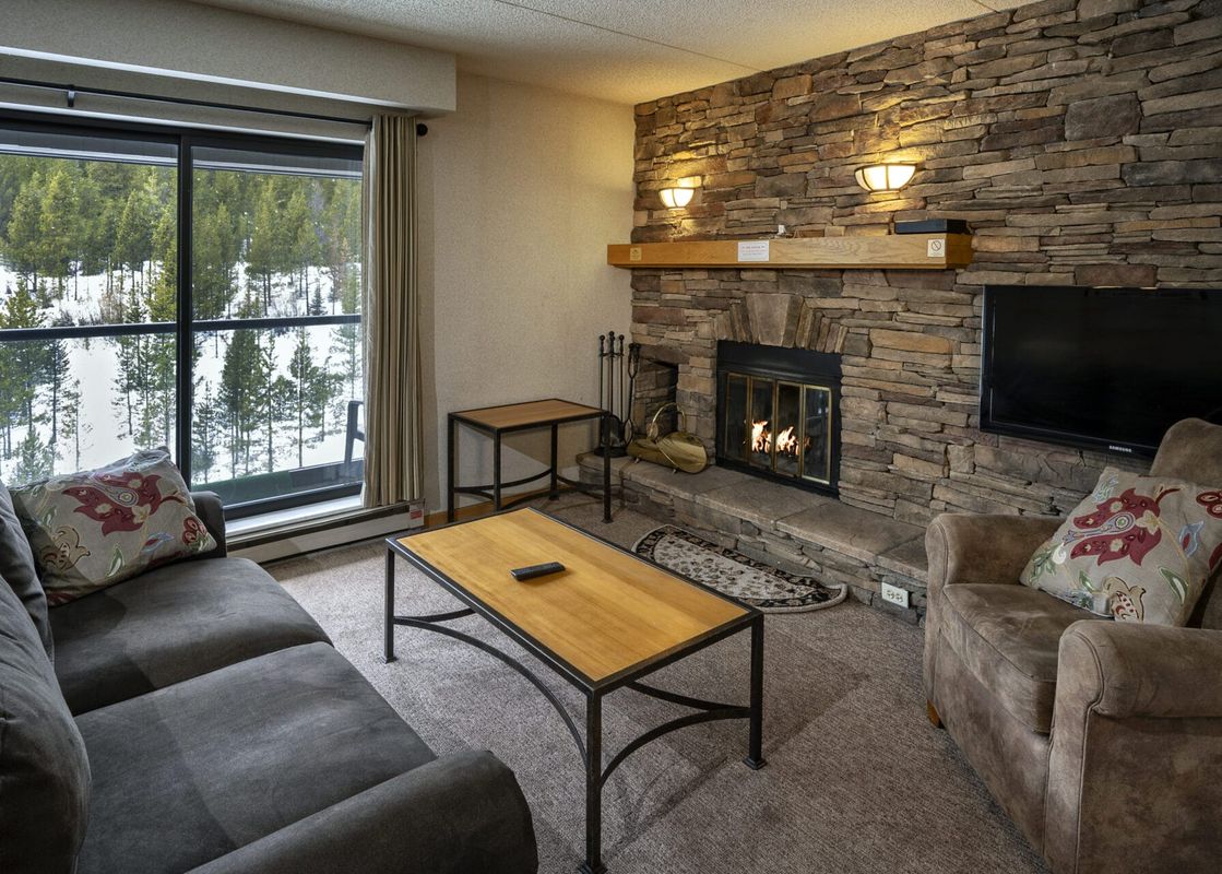 A cozy living room in the Building 4 Two Bedroom Breckenridge Suite features a stone fireplace, comfortable seating, and a large window offering a view of snow-covered pine trees.