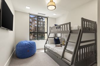 A modern children's bunk room at Horizon View features a gray bunk bed, a blue bean bag, a wall-mounted TV, and a large window with a view of other buildings.