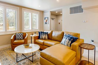 The living area of Meadows Oxford 13 features a tan leather sectional sofa, a matching armchair, a white coffee table, and an area rug, with two windows offering outdoor views.