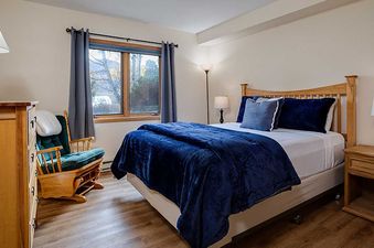 The bedroom at The Pines Condominiums P103C featuring a queen-sized bed with blue bedding, a rocking chair, and natural wood furnishings.