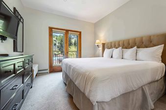 The primary bedroom at The Charter E2305 with a king-sized bed, wall-mounted lamp, dark wood dresser with a television, and French doors opening to a balcony with mountain views.