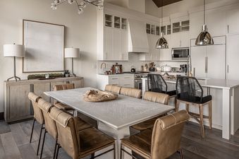 The open concept kitchen and dining area in Inspiration Point Townhome 4C features a long, light wood dining table with eight tan leather chairs, complemented by two black woven bar stools at the kitchen island, all under the glow of modern chandeliers an