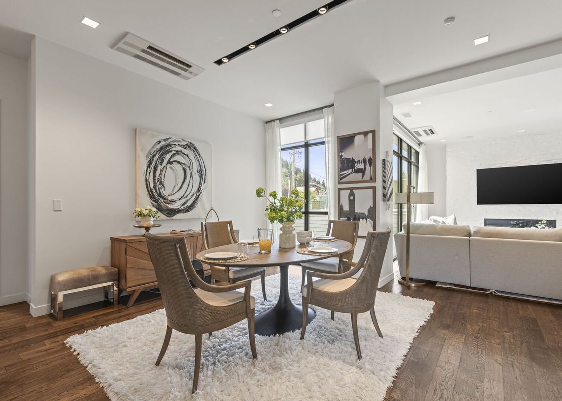 A modern dining area with a round wooden table, four chairs, and a white shag rug, illuminated by natural light and track lighting, with an open-concept lounge area in the background.