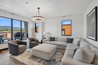 A modern living room at the Grandview Escape with a large sectional sofa, two armchairs, an ottoman, a stylish chandelier, and expansive windows offering mountain views.