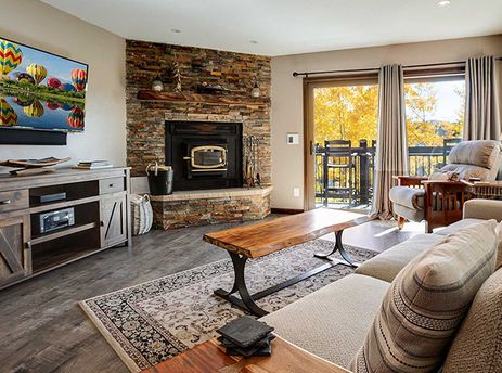 A cozy lounge area with a stone fireplace, large TV, and access to a balcony overlooking autumn trees.