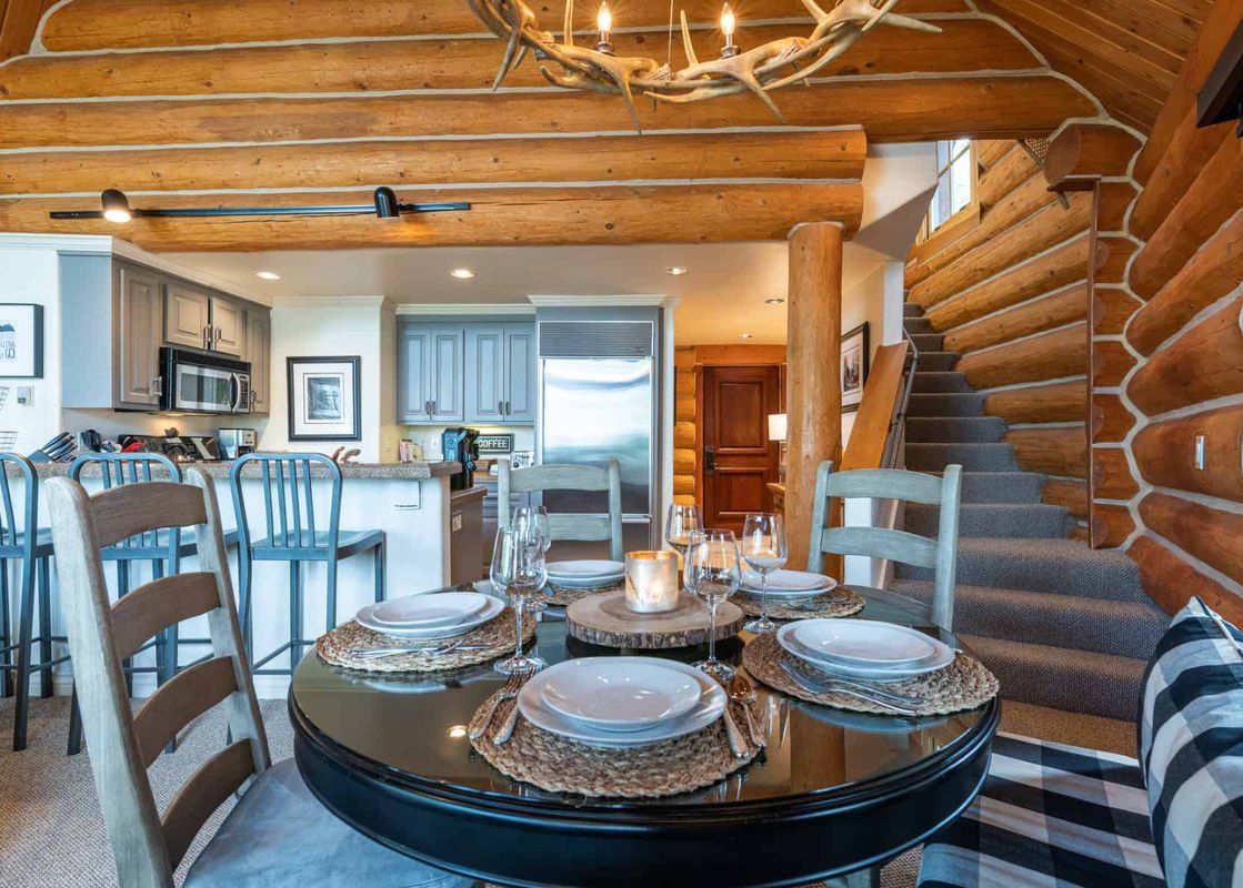 A dining table with four place settings set with plates, cutlery, and glasses, is in the center of the image.  There is a large rustic wooden chandelier above the table.  The table is situated in a log cabin with a stairway leading to a loft. The kitchen
