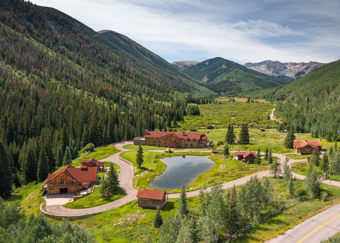 An aerial view of Elk Mountain Lodge, a collection of log buildings with red roofs, nestled in a lush green valley with a pond and winding roads, surrounded by dense pine forests and distant mountain peaks under a partly cloudy sky.