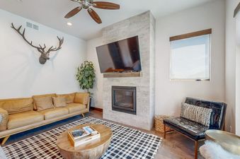 A modern living room featuring a tan leather sofa, a round wooden coffee table, a fireplace with a TV mounted above it, a black leather armchair, and a deer head decor on the wall.
