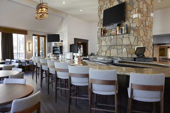 A sophisticated bar area at The Charter F255 features a long granite-topped bar with upholstered stools, a stacked stone column with a TV, and elegant lighting, with dining tables and large windows offering an outdoor view.