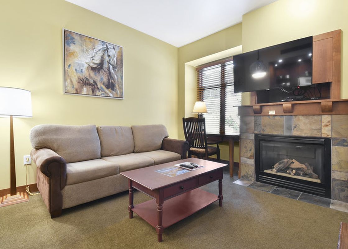 A cozy lounge area in the Sundial Lodge Studio featuring a sofa, a red coffee table, a fireplace, and a television mounted above the mantel.