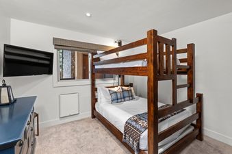 A cozy bunk room at Crestwood 1201 features a wooden bunk bed with white bedding, a wall-mounted TV, a blue dresser, and a window with an outdoor view.