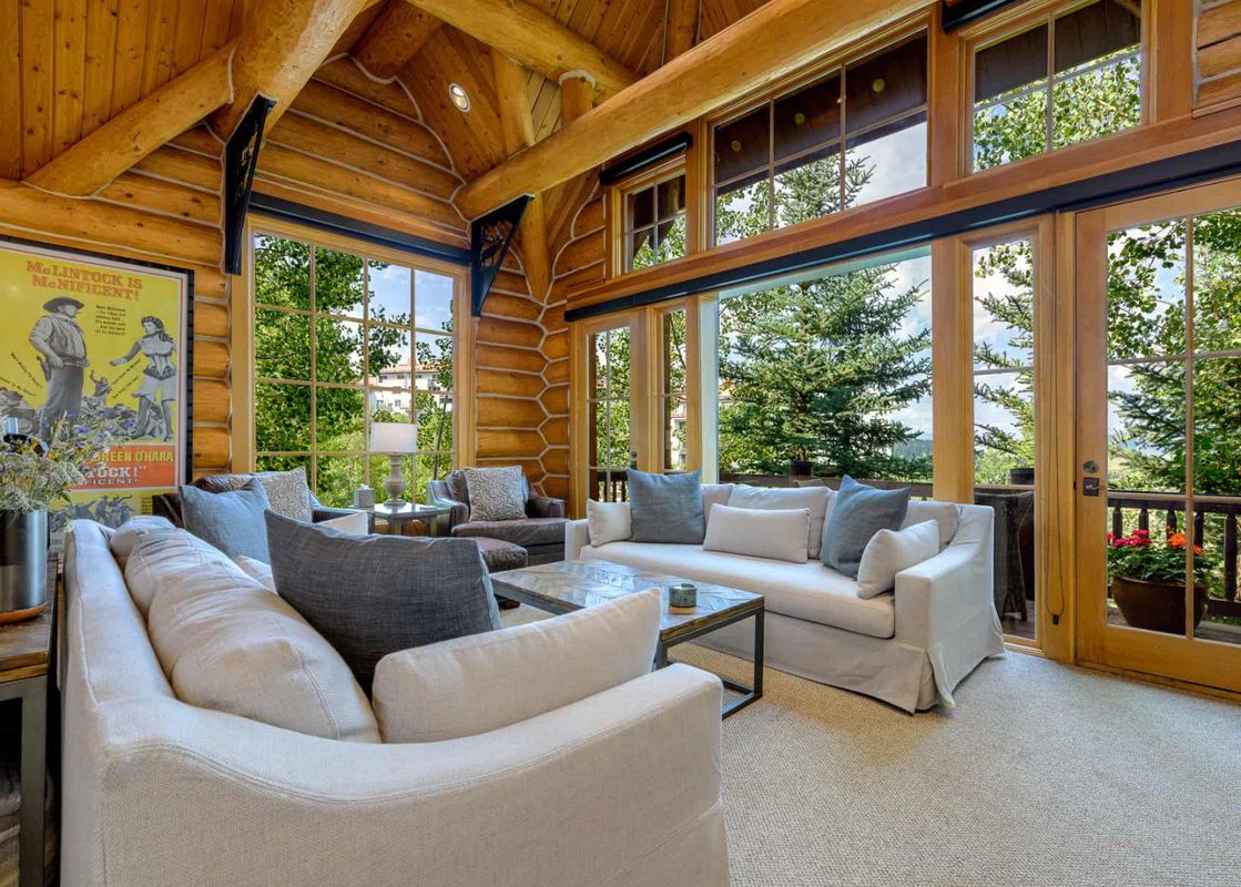 A living room with large windows overlooking a forest, featuring a comfy white couch and two armchairs.
