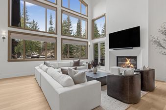A large great room at Red Pine Estate with a white sectional sofa, dark leather armchairs, a fireplace, and a wall of windows offering views of pine trees.
