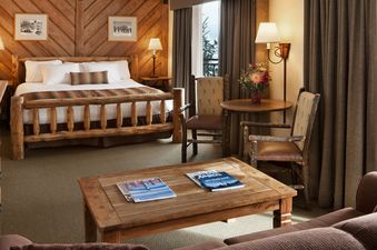 A cozy bedroom in the Stonebridge Inn King Suite, featuring a rustic wooden bed, two armchairs, a coffee table with magazines, and a dining table with chairs, all under warm lighting.