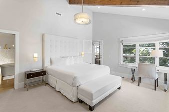 A bright bedroom features a large white bed, desk, and ensuite bathroom.