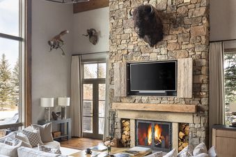 A cozy, rustic-chic living room with a large stone fireplace, a sectional sofa, and a chandelier, featuring large windows with a snowy outdoor view.
