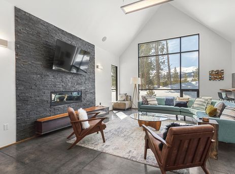 A modern alpine living room featuring a stacked stone fireplace, large windows with mountain views, a teal sectional sofa, and stylish wooden armchairs.
