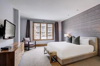 A spacious bedroom in The Arrabelle Four Bedroom GR features a large bed with white linens, a wooden dresser with a TV mounted above it, and a comfortable armchair, all illuminated by natural light from a window with a view of buildings.