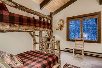A close-up view of the bedroom at Breck Lodge, featuring a unique log bunk bed with red and black buffalo plaid bedding and a window offering a glimpse of the surrounding trees.