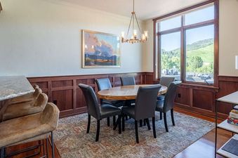 A sophisticated dining area featuring a wooden oval table with six black chairs, an elegant chandelier, a large abstract painting, wooden wainscoting, and a large window with a scenic view of green hills, complemented by a breakfast bar with granite count