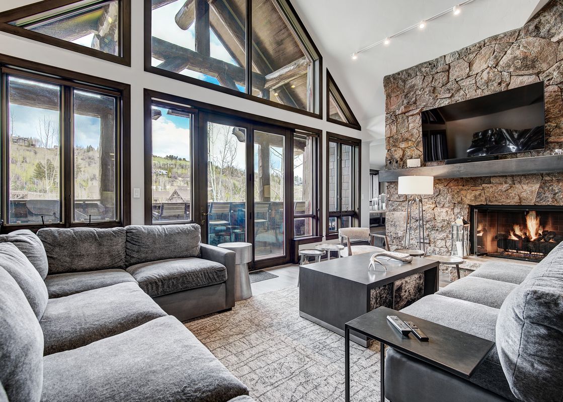 A spacious and inviting living room featuring a large gray sectional sofa, a stone fireplace with a television, and expansive windows offering scenic views of the outdoors.