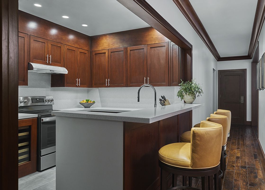 A modern kitchen with rich wood cabinetry, stainless steel appliances, light gray countertops, a sink with a gooseneck faucet, and an island bar with four yellow leather bar stools.