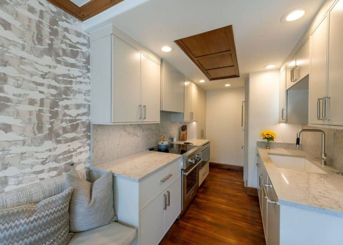 A modern galley-style kitchen in Chateau Beaver Creek Residence 204 features white cabinetry, granite countertops, stainless steel appliances, and a built-in bench with pillows, all illuminated by recessed and under-cabinet lighting.