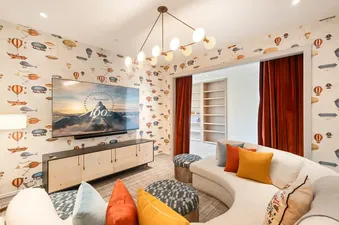 A cozy media room at Galloping Goose Chalet with a curved white sectional sofa, colorful pillows, playful hot-air balloon wallpaper, and a large wall-mounted TV.