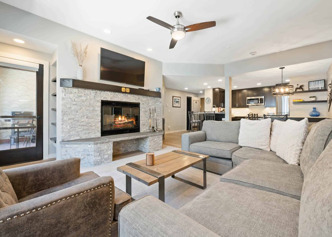 The living room of The Charter A2505 features a comfortable sectional sofa, an elegant armchair, a fireplace with a large TV mounted above it, and an open-concept layout leading into the kitchen.