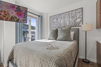 The comfortable bedroom in Galleria 307 features a large bed with a textured comforter, flanked by a scenic window and a wall-mounted TV, with a floor lamp and cabinet on the side.