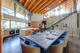An expansive open-concept room featuring a modern kitchen with wooden cabinetry and a large island, adjacent to a dining area with a long wooden table and blue upholstered chairs, all beneath a vaulted wooden ceiling with unique pendant lights.