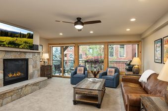 A cozy living room in Montaneros 220 with a stone fireplace, large television, comfortable seating, and a balcony with an outdoor view.