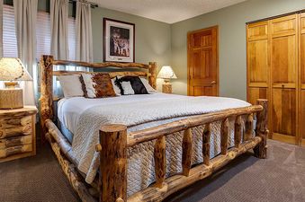 A rustic bedroom at Ranch at Steamboat RA501 with a log-frame bed, white duvet, and cowhide pillows, alongside a wooden nightstand and lamp.