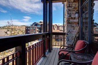A balcony with a view of the surrounding mountains and other condos. The balcony has two chairs and a table, and there is a stone wall in the background.