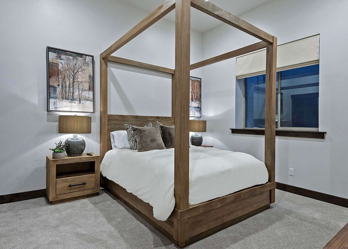 A luxurious bedroom at the Retreat at Black Rock Ridge features a large wooden canopy bed with white linens and gray pillows, flanked by two wooden nightstands with lamps and framed artwork, illuminated by natural light from two windows.