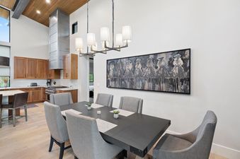A modern dining area with a long dark table and six gray upholstered chairs, beneath a sleek chandelier, with an open-concept kitchen featuring wooden cabinets and stainless steel appliances in the background.