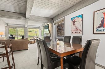 A sophisticated dining area featuring a polished wooden table with a glass top and six black leather chairs, set against a backdrop of large windows offering views of green hills and walls adorned with vintage ski posters.