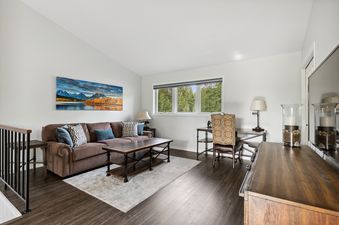 A spacious living room at Tee Time featuring a brown sofa, a wooden coffee table, and a desk, all set against dark hardwood floors and white walls, with a large window revealing a view of green trees.