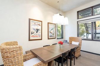 A dining room with a wooden table and chairs, a white ceiling, and a large window with a view of trees.