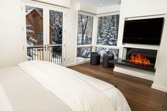 A modern bedroom with a king-sized bed, a wall-mounted TV, a fireplace, and large windows offering views of a snow-covered landscape and a private balcony.