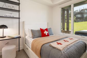 A bedroom at Moonlight Basin 9 Wildwood, featuring a plush bed with a tan blanket, a Swiss flag throw, and large windows looking out onto the scenic surroundings.