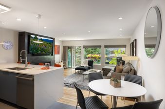 A modern open-concept living area in Montaneros 214 featuring a kitchen island, dining table, and a comfortable living room with a TV and balcony view.