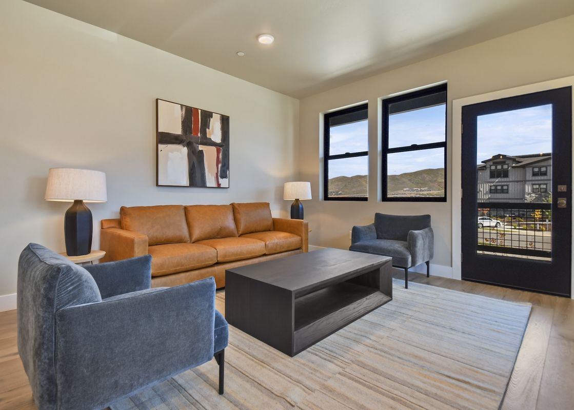 A modern living area featuring a tan leather sofa, gray armchairs, a dark wood coffee table, and an abstract painting, with large windows and a glass door providing views of the exterior.