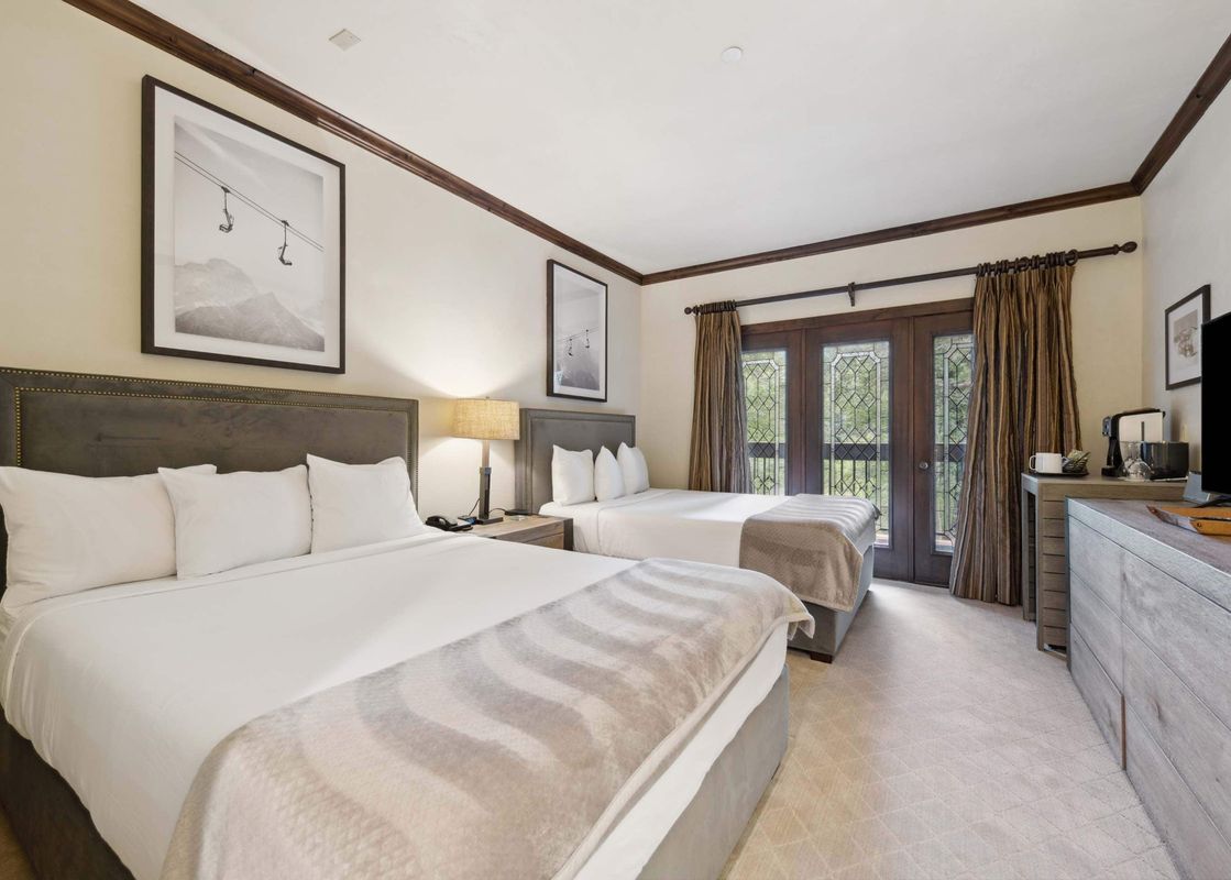 A luxurious twin queen bedroom at The Charter C115 features two plush beds with white linens, framed ski lift artwork, dark wooden french doors leading to a balcony, a chic bedside lamp, and a dresser with a flat-screen TV.