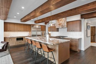 A modern kitchen with a large marble-topped island, wooden cabinetry, stainless steel appliances, and leather bar stools under exposed wooden beams and stylish pendant lights.